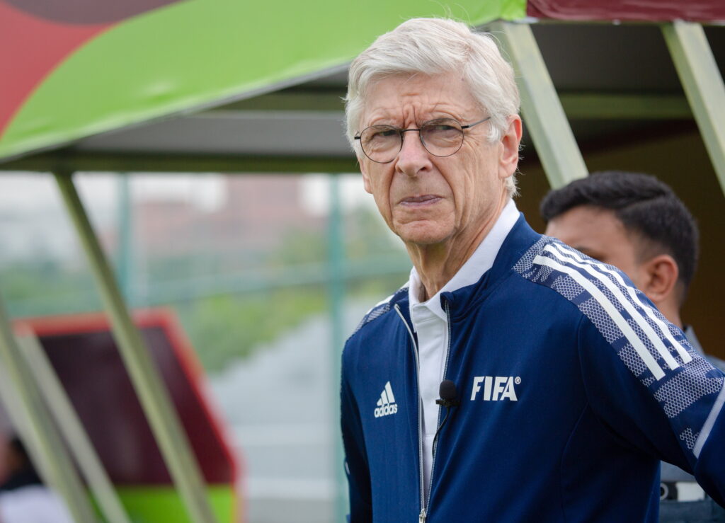 Arsène Wenger wearing glasses and a blue FIFA tracksuit jacket.