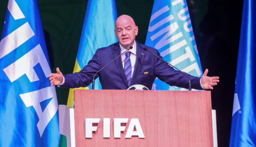 Gianni Infantino speaks behind a FIFA-branded podium with his arms outstretched and flags behind him.