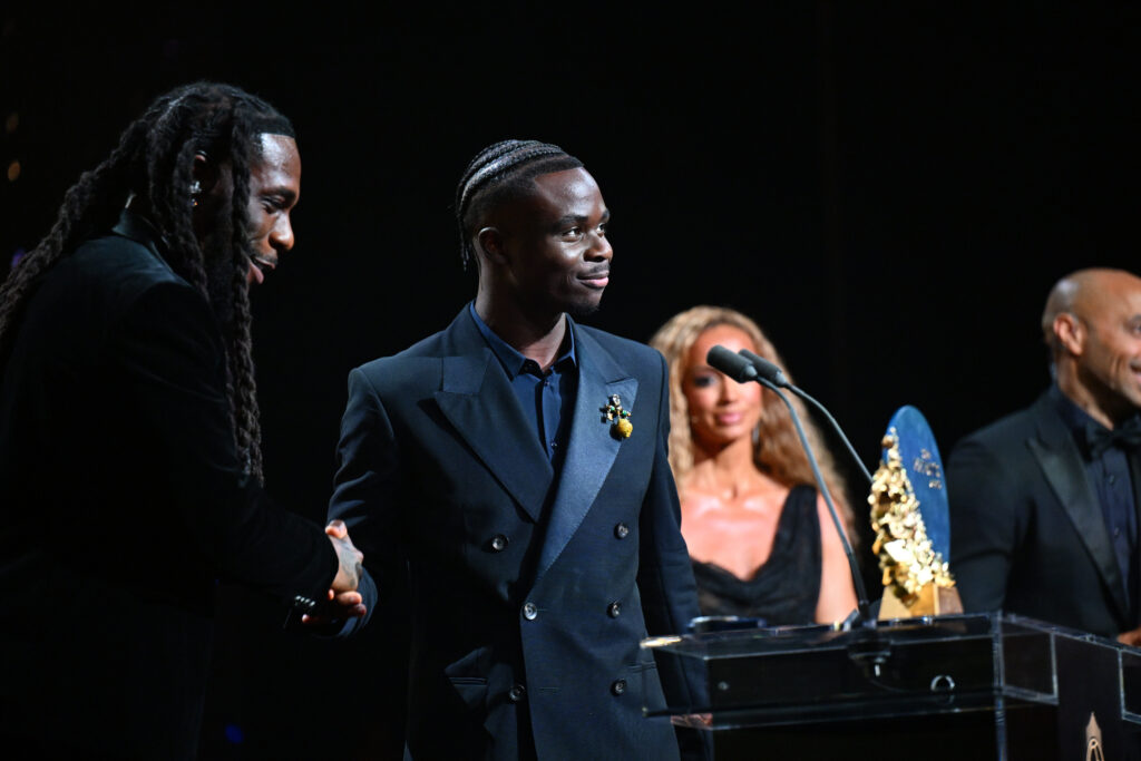 Bukayo Saka shakes hands with Burna Boy beside the Sócrates Award trophy at the 2026 Ballon d’Or ceremony.